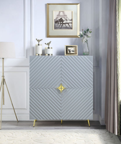 A modern room featuring the Gaines High Gloss Accent Cabinet in gray with gold accents and gold legs. Decorative items, a flower vase, and framed photos sit on top. A floor lamp and curtains flank each side.