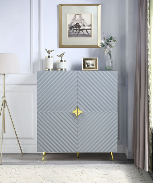 A modern room featuring the Gaines High Gloss Accent Cabinet in gray with gold accents and gold legs. Decorative items, a flower vase, and framed photos sit on top. A floor lamp and curtains flank each side.