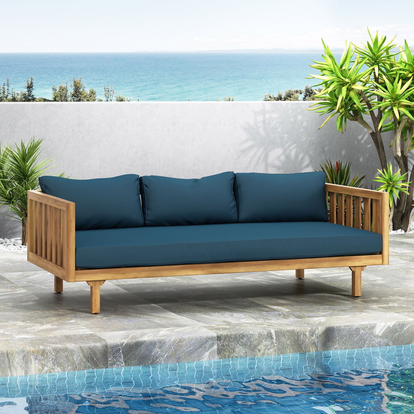 The Claremont 3 Seater Daybed, made of teak acacia wood with water-resistant cushions, is placed on a stone patio by the pool, surrounded by greenery with ocean and sky views in the background.