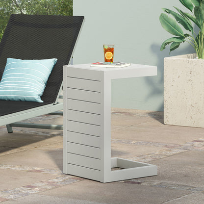 The Aluminum Outdoor C-Shaped Side Table for Patio holds a glass of iced tea with lemon and orange slices. Next to it is a black lounge chair with a blue-striped pillow, and a large potted plant sits nearby.