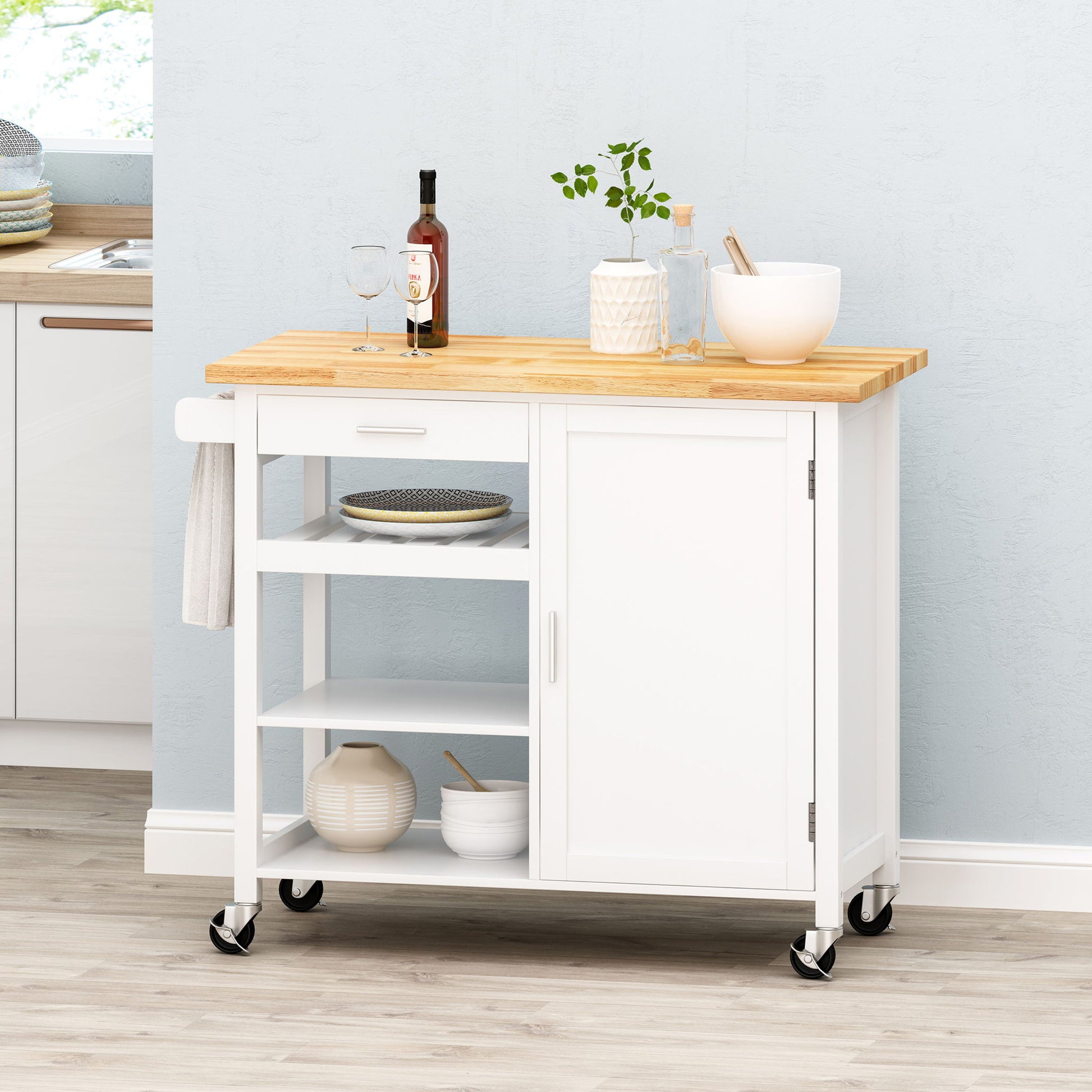 The Contemporary Design Kitchen Cart With Wheels features a white frame, wooden top, open shelves, drawer, cabinet, and towel rack. Displayed in a bright kitchen with wine, glasses, a bowl, tableware, and a plant on top.