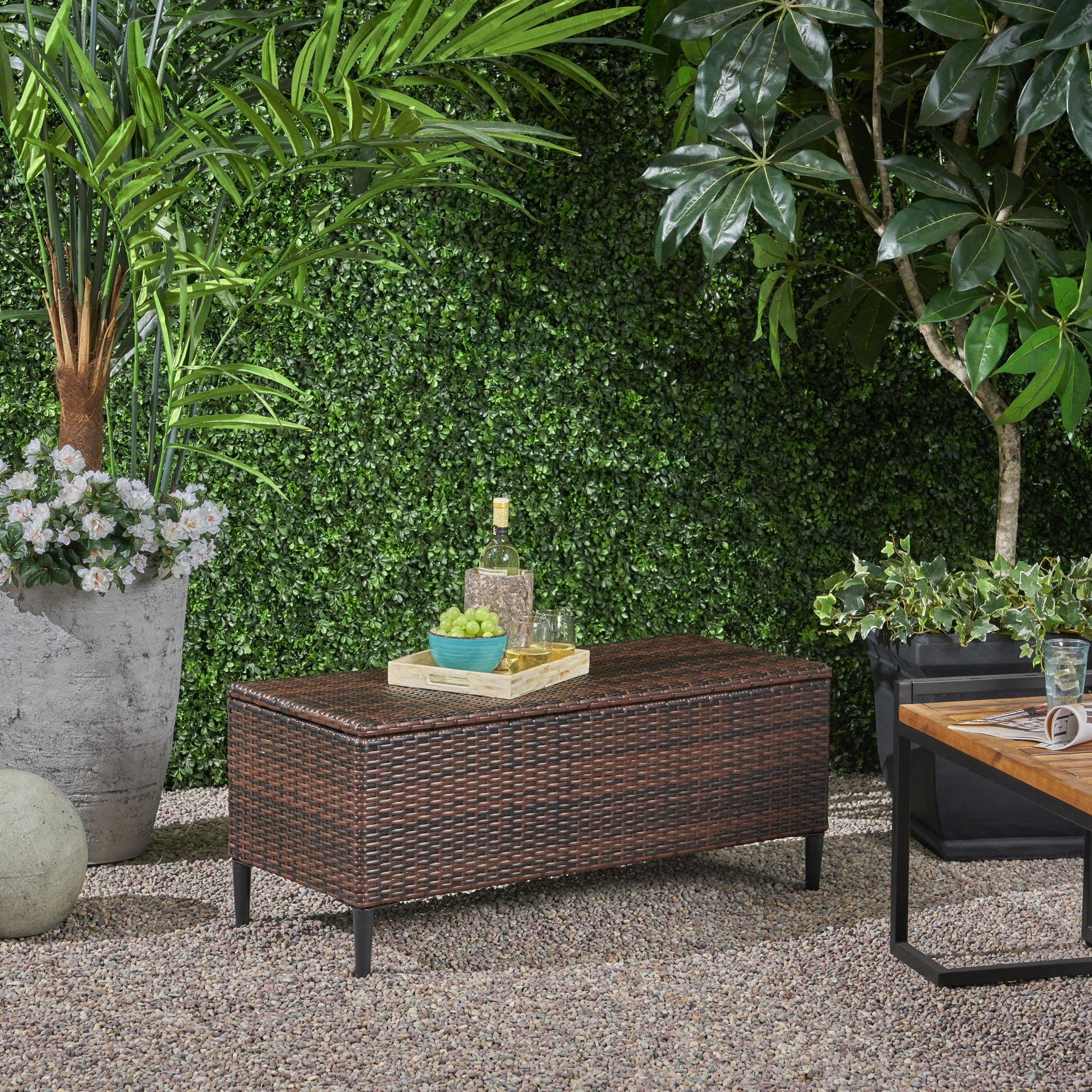 An outdoor patio features the Rupert - Storage Unit Resin Wicker Outdoor bench topped with a tray holding wine and snacks, surrounded by lush greenery and a small wooden table on gravel.