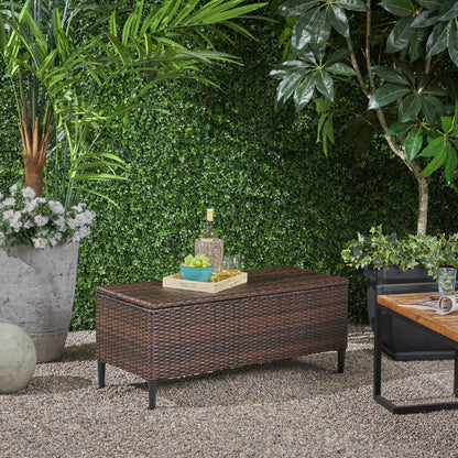 An outdoor patio features the Rupert - Storage Unit Resin Wicker Outdoor bench topped with a tray holding wine and snacks, surrounded by lush greenery and a small wooden table on gravel.
