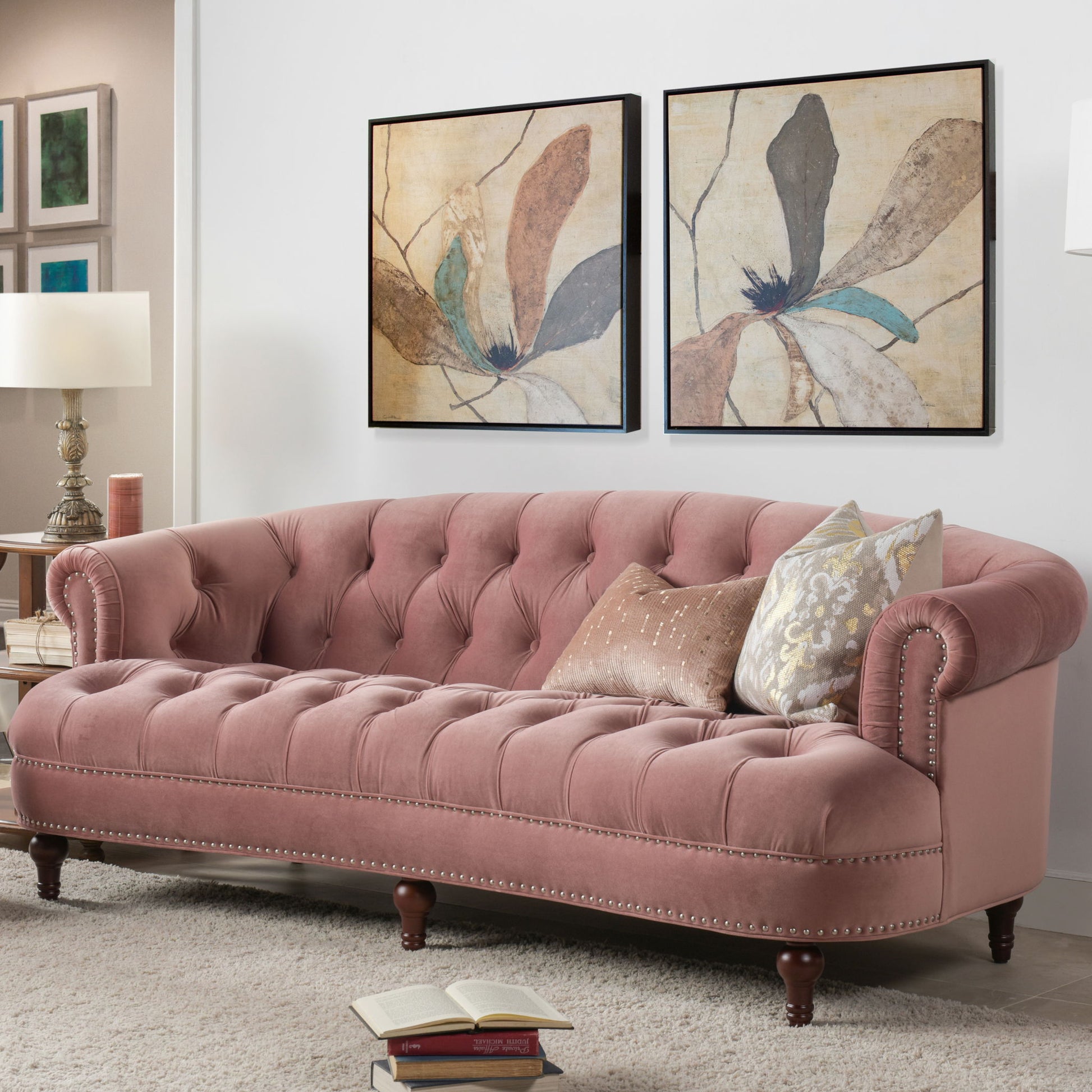 The La Rosa Victorian Chesterfield Tufted Sofa in pink sits against a white wall beneath two floral art prints. Patterned accent pillows, an open stack of books on the floor, and a lamp with framed art complete the scene.