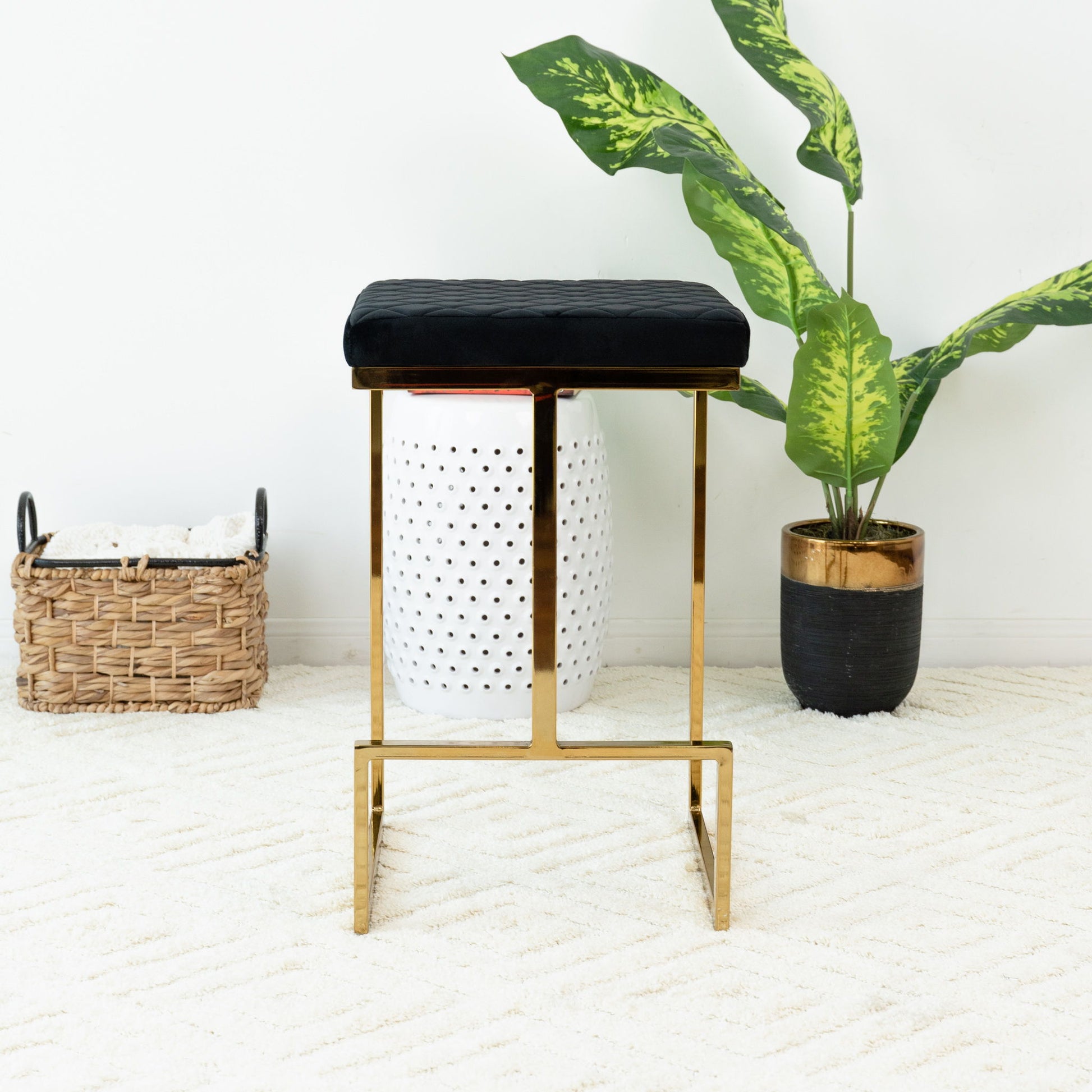 The Joel - Mid Century Modern Luxury Upholstered Stool, featuring a gold metal frame and black cushioned seat, sits on a white textured rug beside a potted green plant and woven basket in a bright, minimalist room.