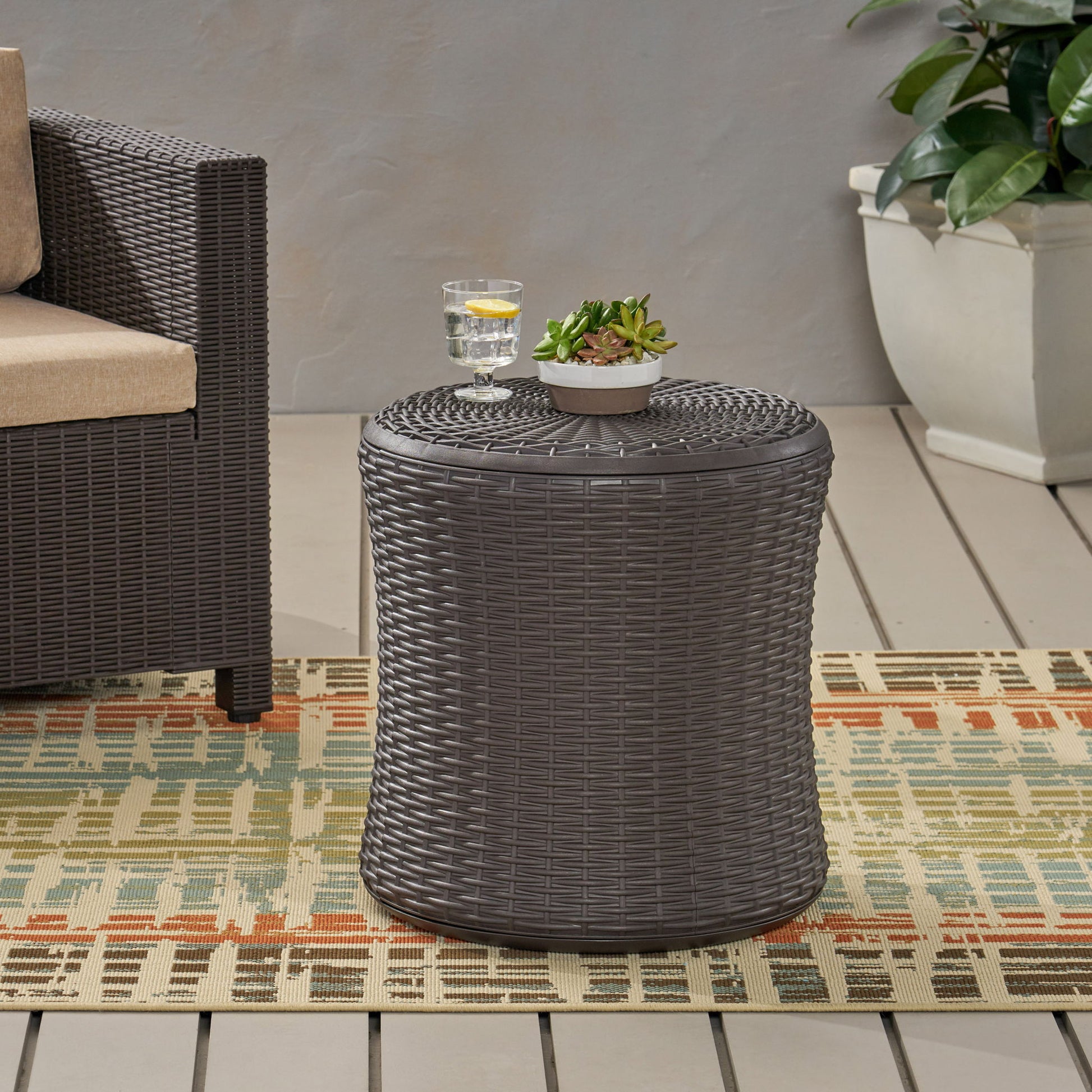 The Olivo - Patio Table Outdoor Side Table in dark brown holds a small potted succulent and a glass of water. It's placed on a patterned rug next to a wicker chair and large potted plant on the patio.
