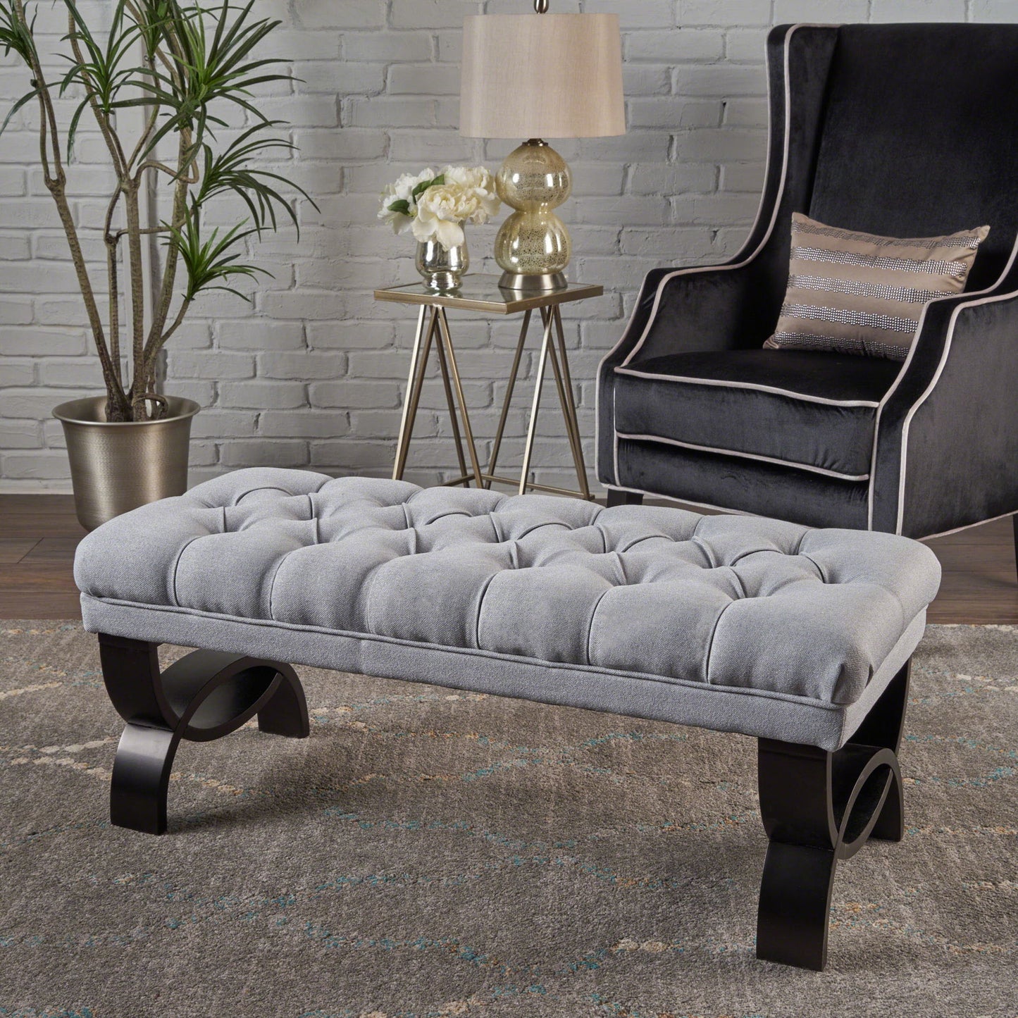 The Hilton - Ottoman Bench Classic Style, tufted in gray with dark curved legs, is on a rug before a gray wingback chair. Nearby are a side table with a lamp, flowers, and décor, plus a potted plant in the corner by the white brick wall.