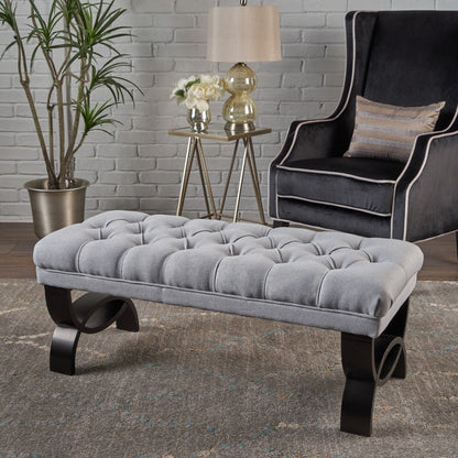 The Hilton - Ottoman Bench Classic Style, tufted in gray with dark curved legs, is on a rug before a gray wingback chair. Nearby are a side table with a lamp, flowers, and décor, plus a potted plant in the corner by the white brick wall.
