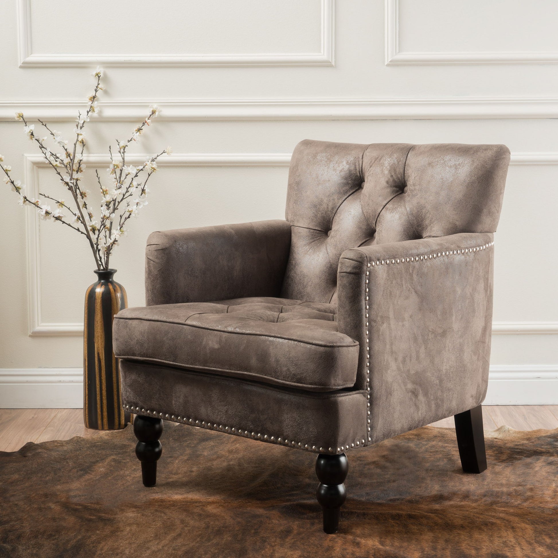 The Vitality Club Chair, featuring button tufted diamond stitching and turned legs in gray, sits on a brown rug beside a tall vase with white flowers against a cream-paneled wall.