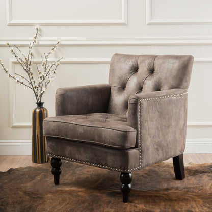 The Vitality Club Chair, featuring button tufted diamond stitching and turned legs in gray, sits on a brown rug beside a tall vase with white flowers against a cream-paneled wall.