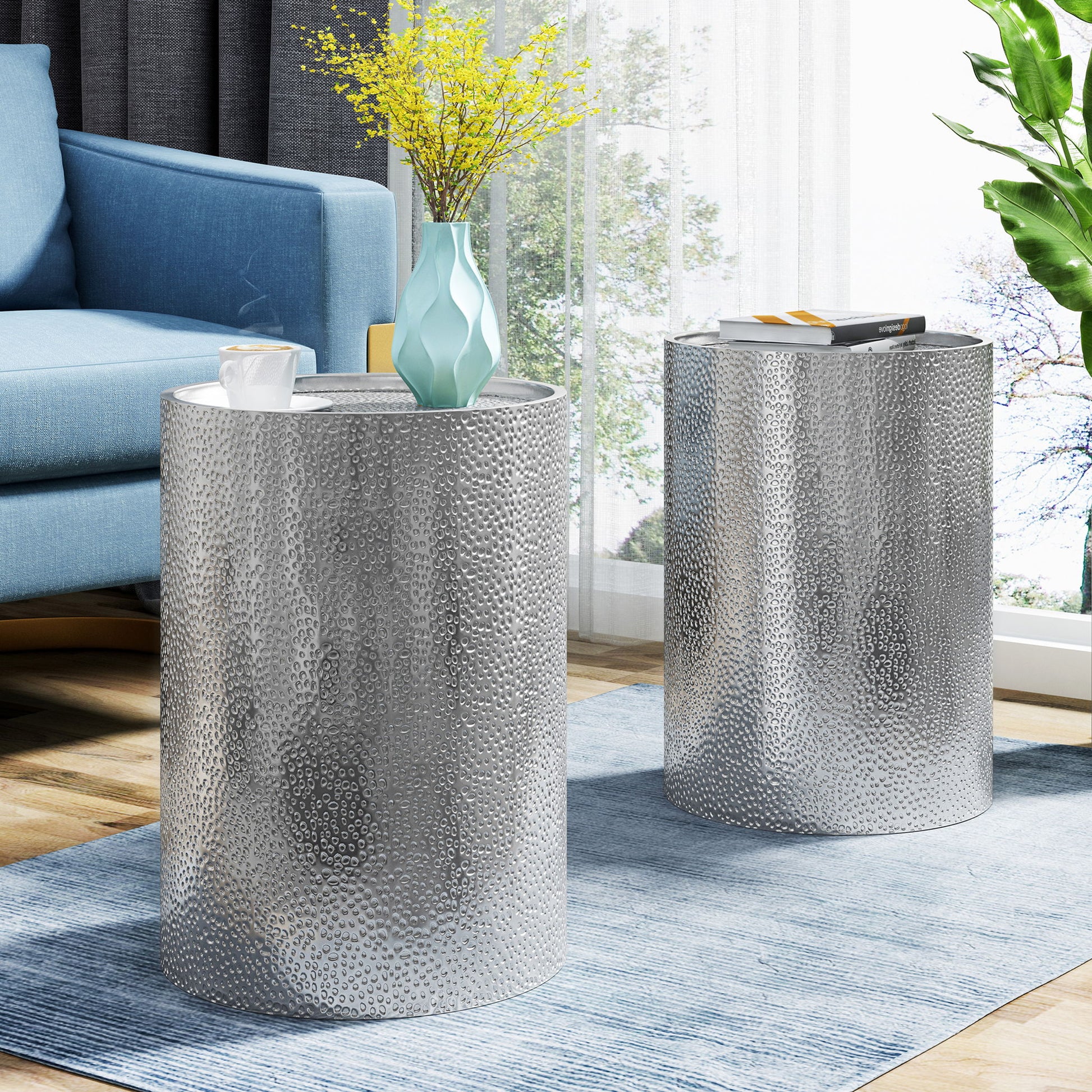 Two Modern Round Accent Tables with hammered surfaces sit on a blue rug beside a blue sofa. One table holds a blue vase with yellow flowers and a cup, the other has a book. Sunlight floods in from large windows.