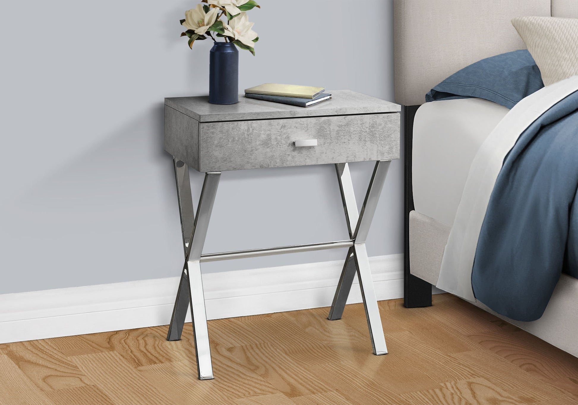 The Accent Side X Table with a storage drawer and metallic legs stands by the bed. On it are a navy vase with white flowers and two closed books. The room features a wooden floor and light gray walls for a modern touch.