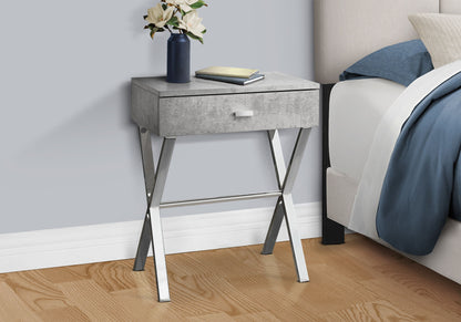 The Accent Side X Table with a storage drawer and metallic legs stands by the bed. On it are a navy vase with white flowers and two closed books. The room features a wooden floor and light gray walls for a modern touch.