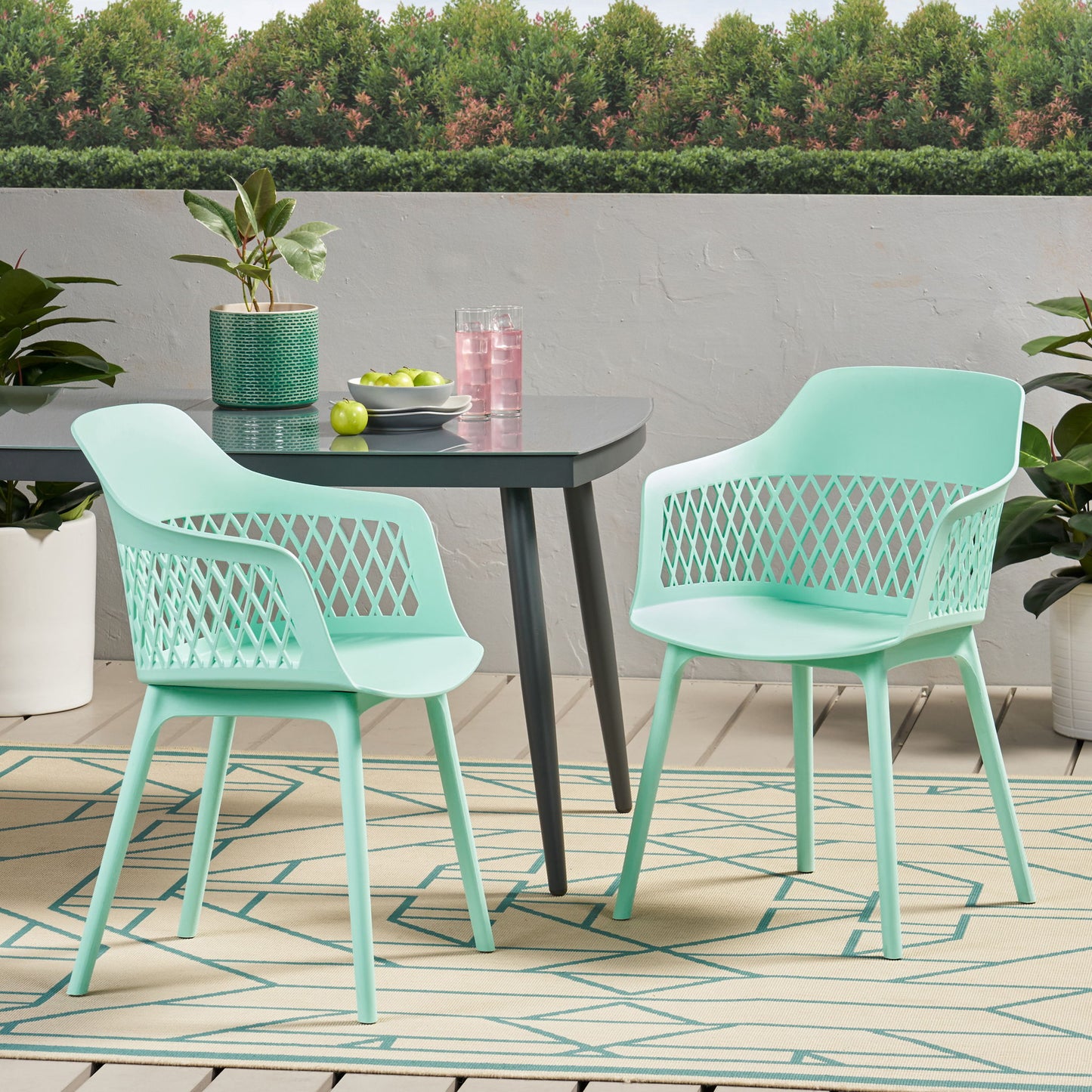 Two Azalea mint green chairs and a dark table with plants, pink drink pitcher, glasses, and green apples on a patterned rug on the patio, surrounded by greenery and trees.