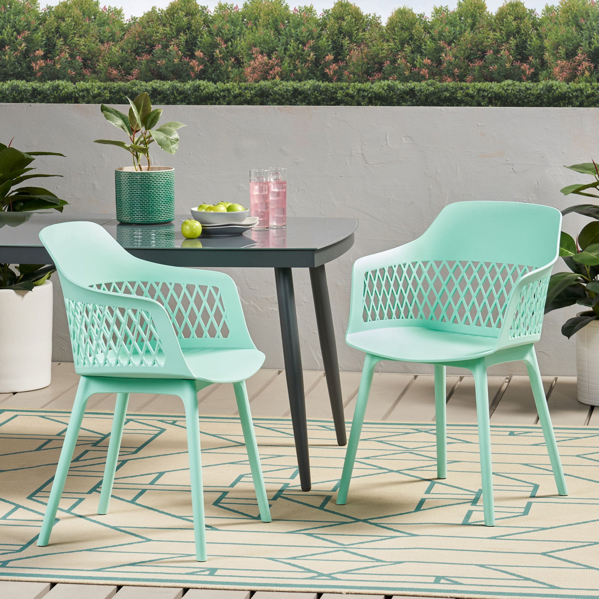 Two Azalea mint green chairs and a dark table with plants, pink drink pitcher, glasses, and green apples on a patterned rug on the patio, surrounded by greenery and trees.
