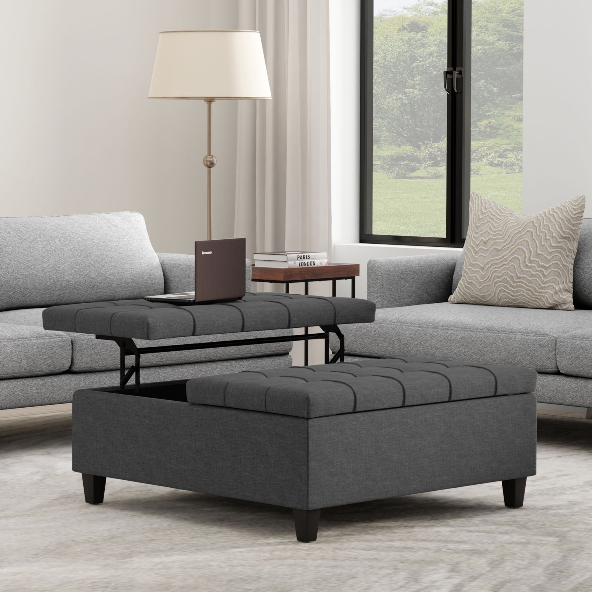 A modern living room with the Harrison Upholstered Coffee Table Storage Ottoman featuring a lift-top compartment, surrounded by a gray sofa and loveseat, plus a round side table with a book, pillow, lamp, and a large window in the background.