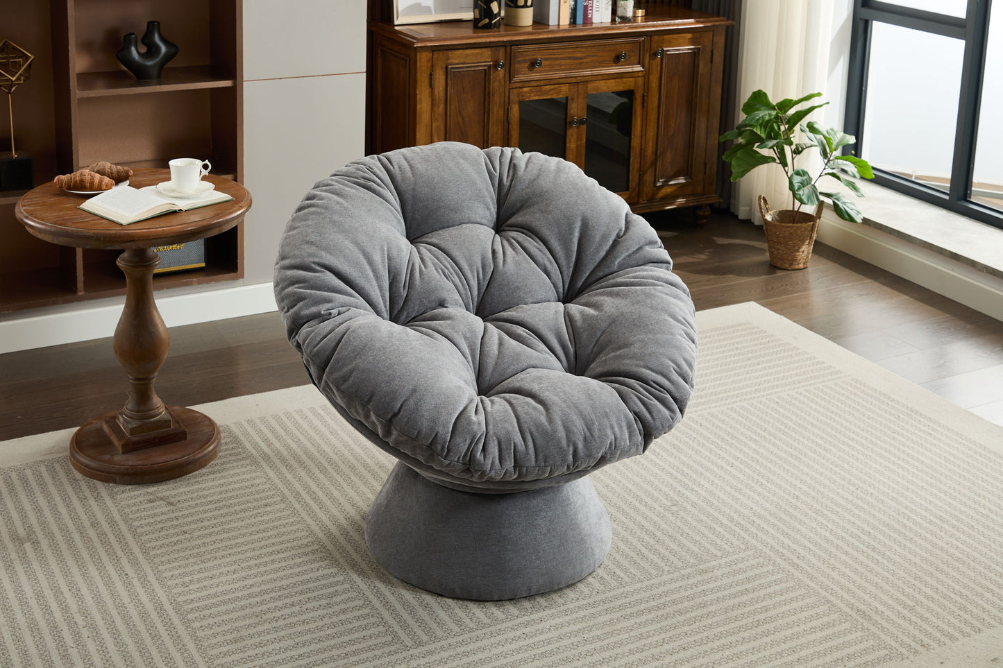 An Oversized Swivel Accent Chair with tufted cushions sits on a patterned rug in a bright living room, near a wooden side table and potted plant by the window. This 360 swivel barrel papasan chair suits living rooms and bedrooms.