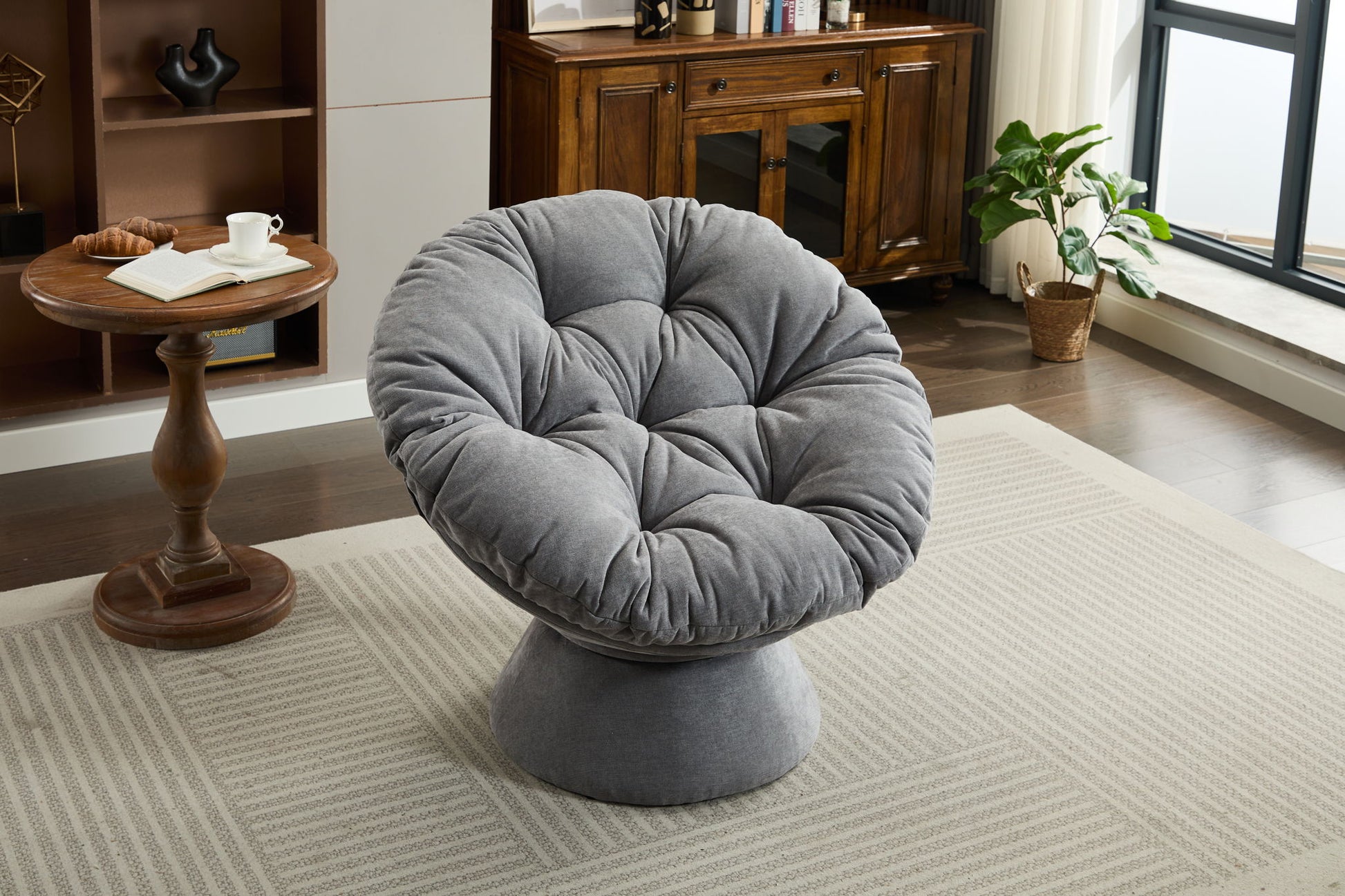 An Oversized Swivel Accent Chair with tufted cushions sits on a patterned rug in a bright living room, near a wooden side table and potted plant by the window. This 360 swivel barrel papasan chair suits living rooms and bedrooms.