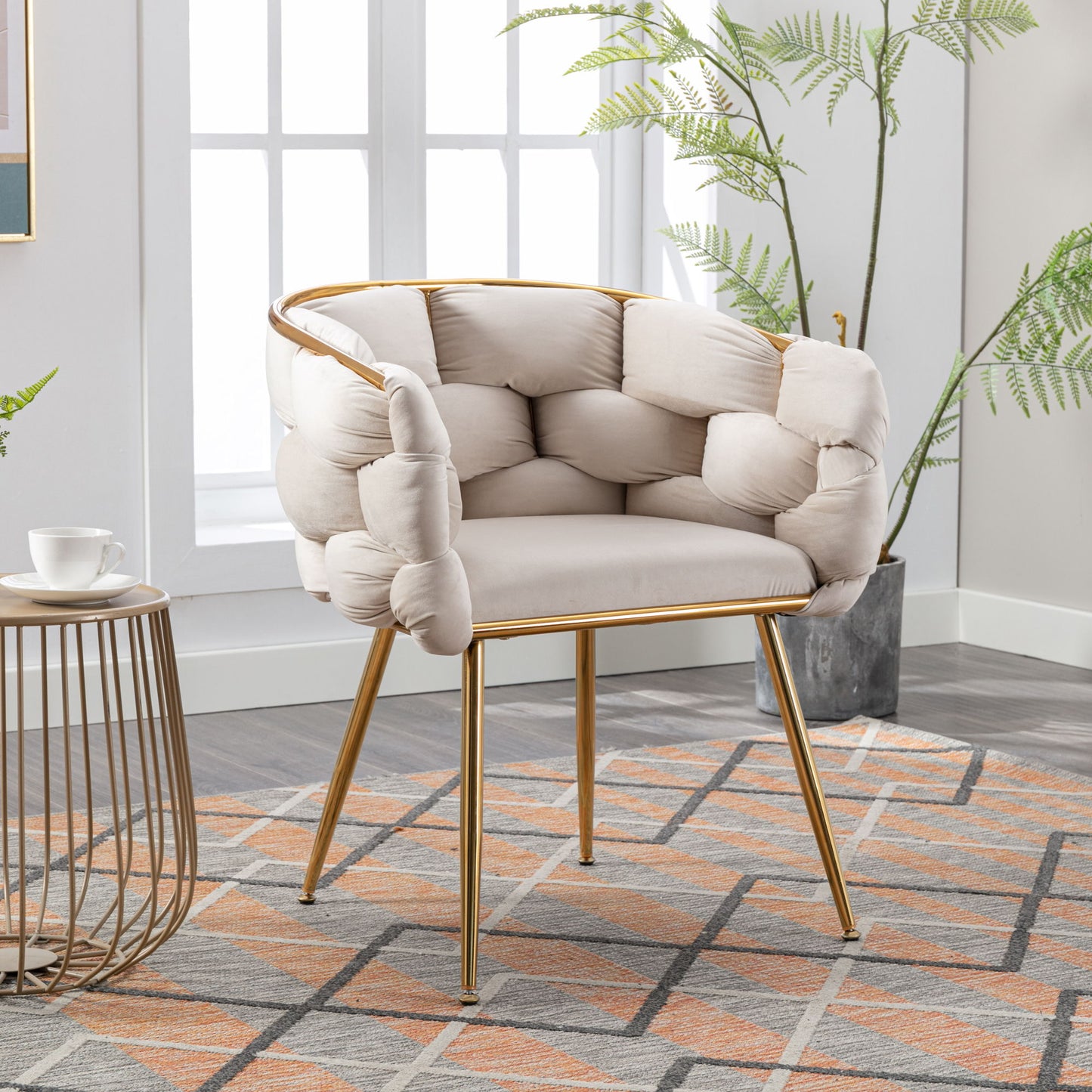 The Luxury Modern Simple Leisure Velvet Single Sofa Chair, featuring a tufted pillow-like backrest and gold metal legs, sits on a patterned rug near a small round side table with a cup in a bright, plant-filled room.