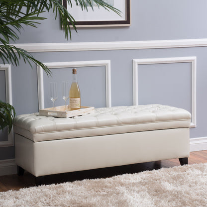 The Juliana Storage Ottoman with Birch Legs sits against a light gray wall with decorative molding. On top, a tray holds a champagne bottle and two flutes. Nearby are a potted plant and a beige rug.