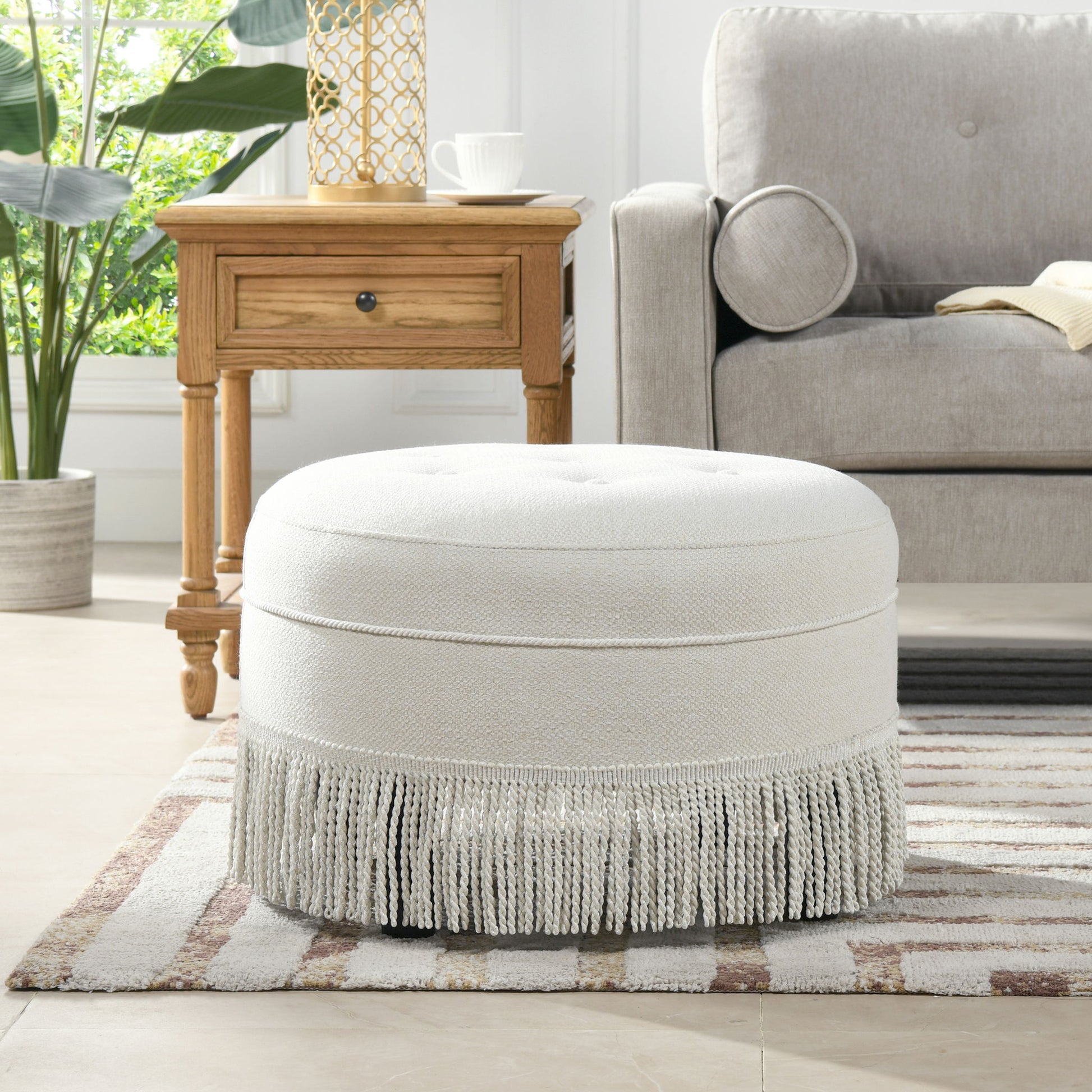 The Yolanda Round Upholstered Accent Ottoman, in light beige with fringe trim, sits on a striped rug next to a wooden side table and light gray sofa in a bright living room filled with plants.