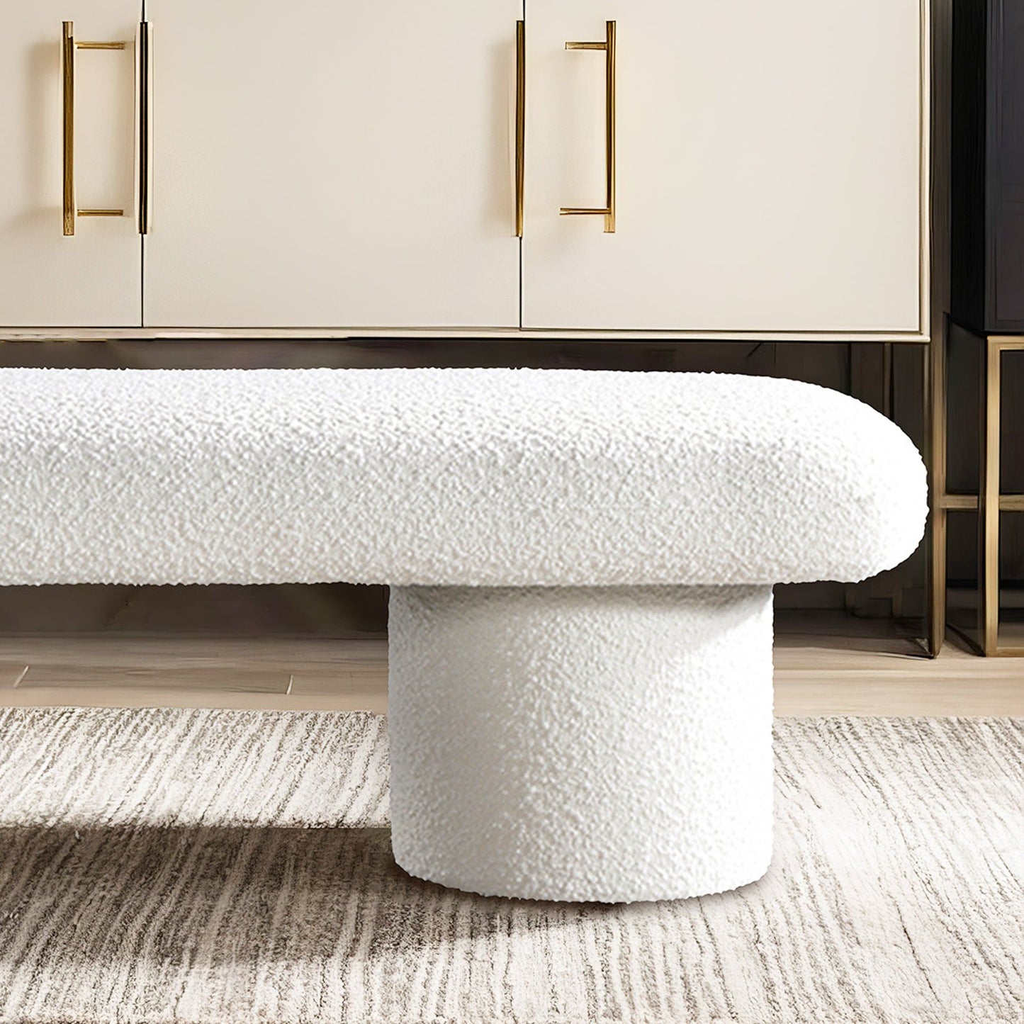 The Randolph - Bench - Ivory features a modern white textured design with a cylindrical base, shown on a light rug in front of a cabinet with gold handles.