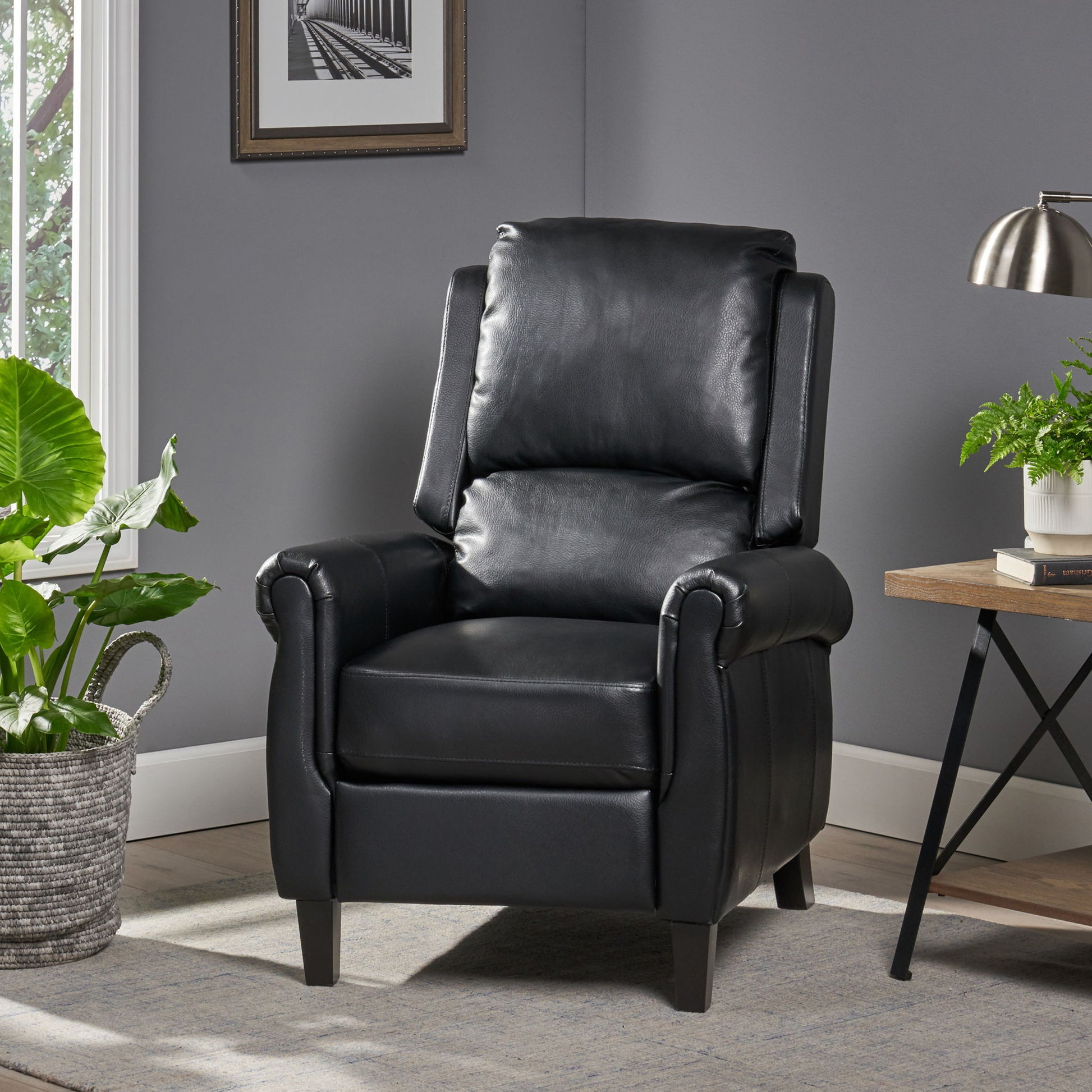 A Contemporary Push Back Recliner Chair in black leather is placed in a modern living room with gray walls, framed artwork, a large potted plant, a wooden desk with lamp and greenery, and natural light streaming through the window.
