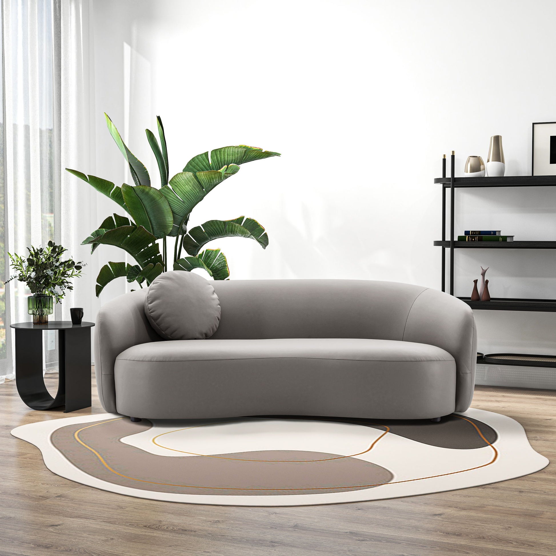 A modern living room featuring the Porter - Comfortable Sofa in gray, a round accent pillow, abstract-patterned rug, potted plant, black side table with vase, and black shelving against a white wall with wood flooring and natural light.