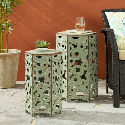 The Selva Accent Table Set, in sage green metal with geometric cut-outs, is arranged on a stone patio beside a wicker chair and potted plant—one table holds wine and a book, the other displays a succulent.