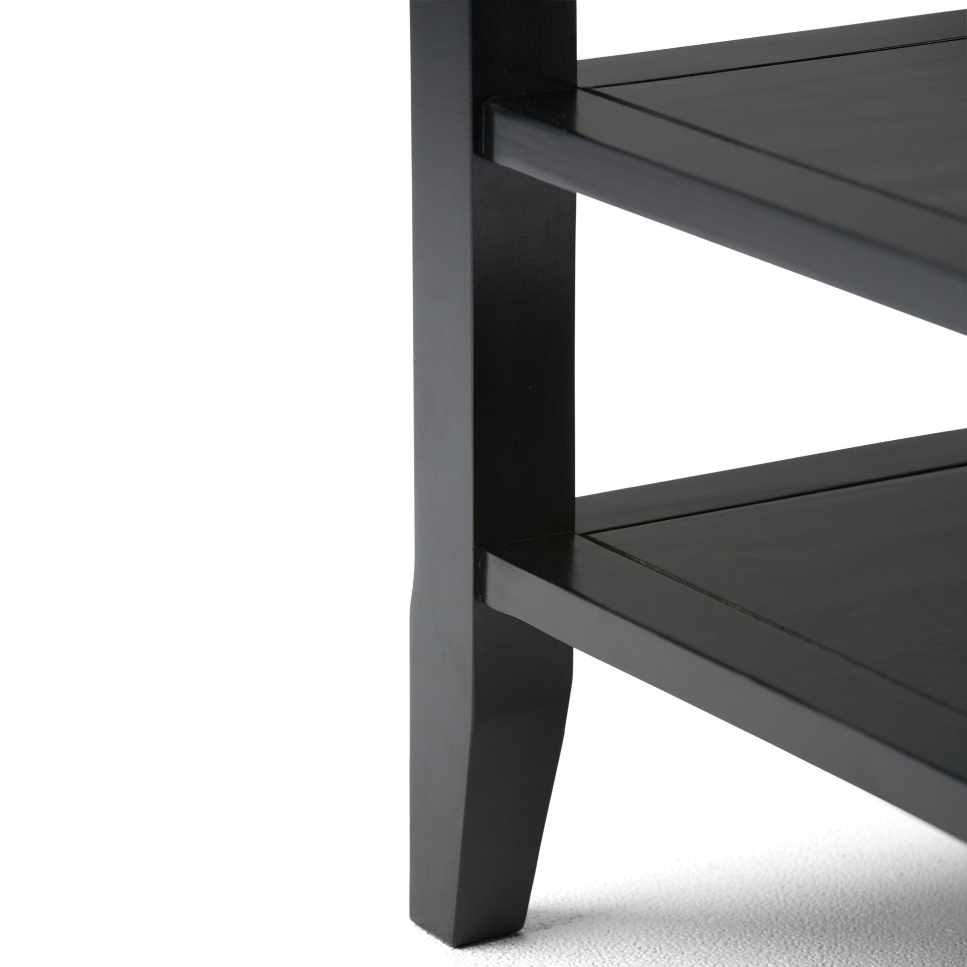 Close-up of the lower corner of the Acadian - Handcrafted End Table in black wood, featuring two shelves and one leg on a light-colored surface.
