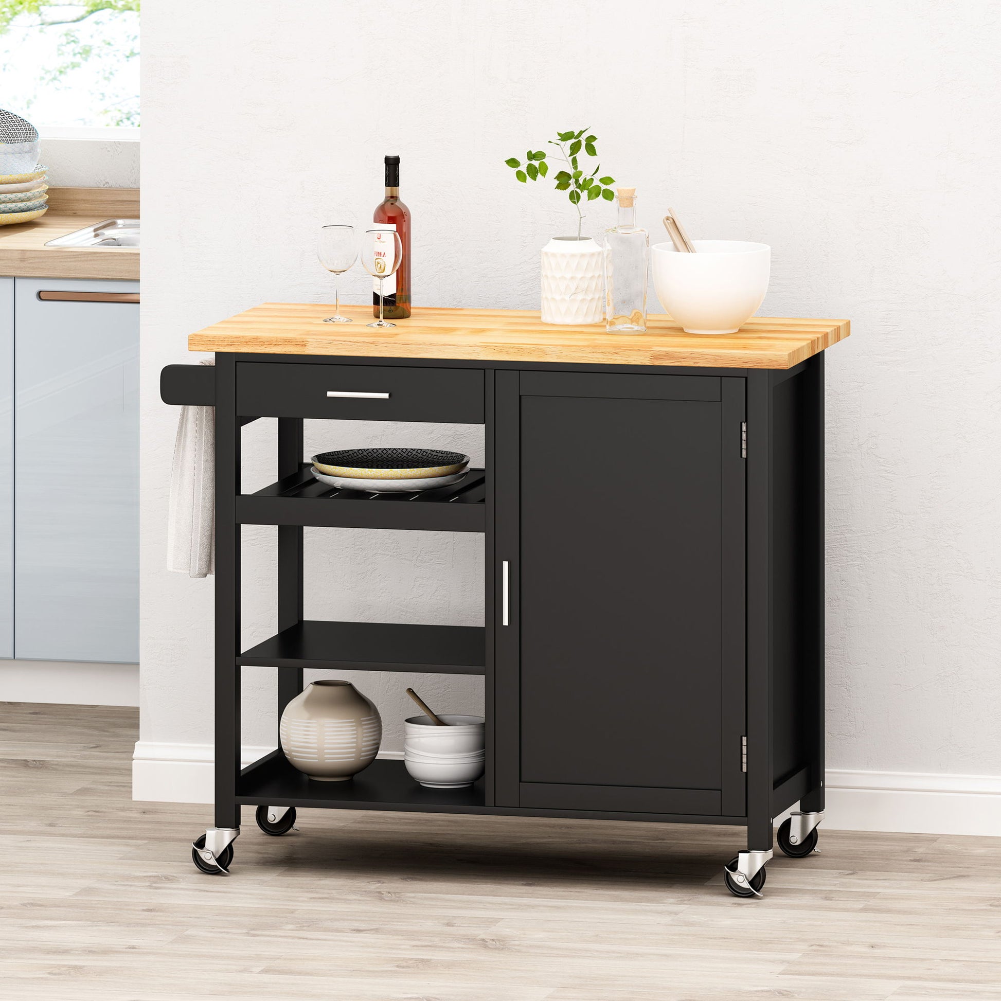 The Contemporary Design Kitchen Cart With Wheels features a black frame, wooden top, open shelves, a drawer, cabinet, and side handle. It holds dishes, wine bottle, two glasses, plant, bowl, and cooking utensils.