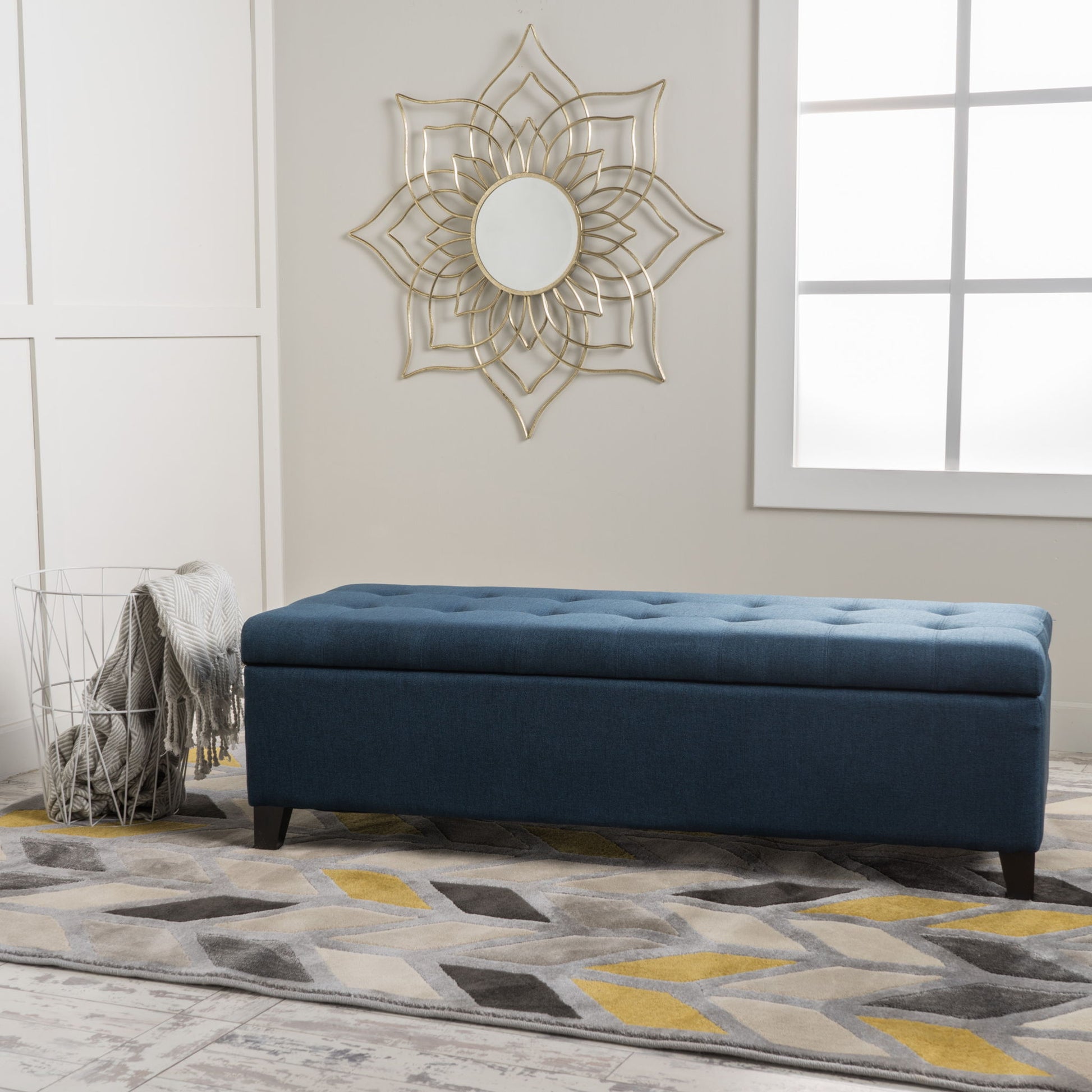 The Nebulous Navy Blue Upholstered Ottoman with storage and a diamond-tufted seat is placed on a geometric-patterned rug, beside a wire basket with a gray throw and below a gold sunburst mirror in this modern room.