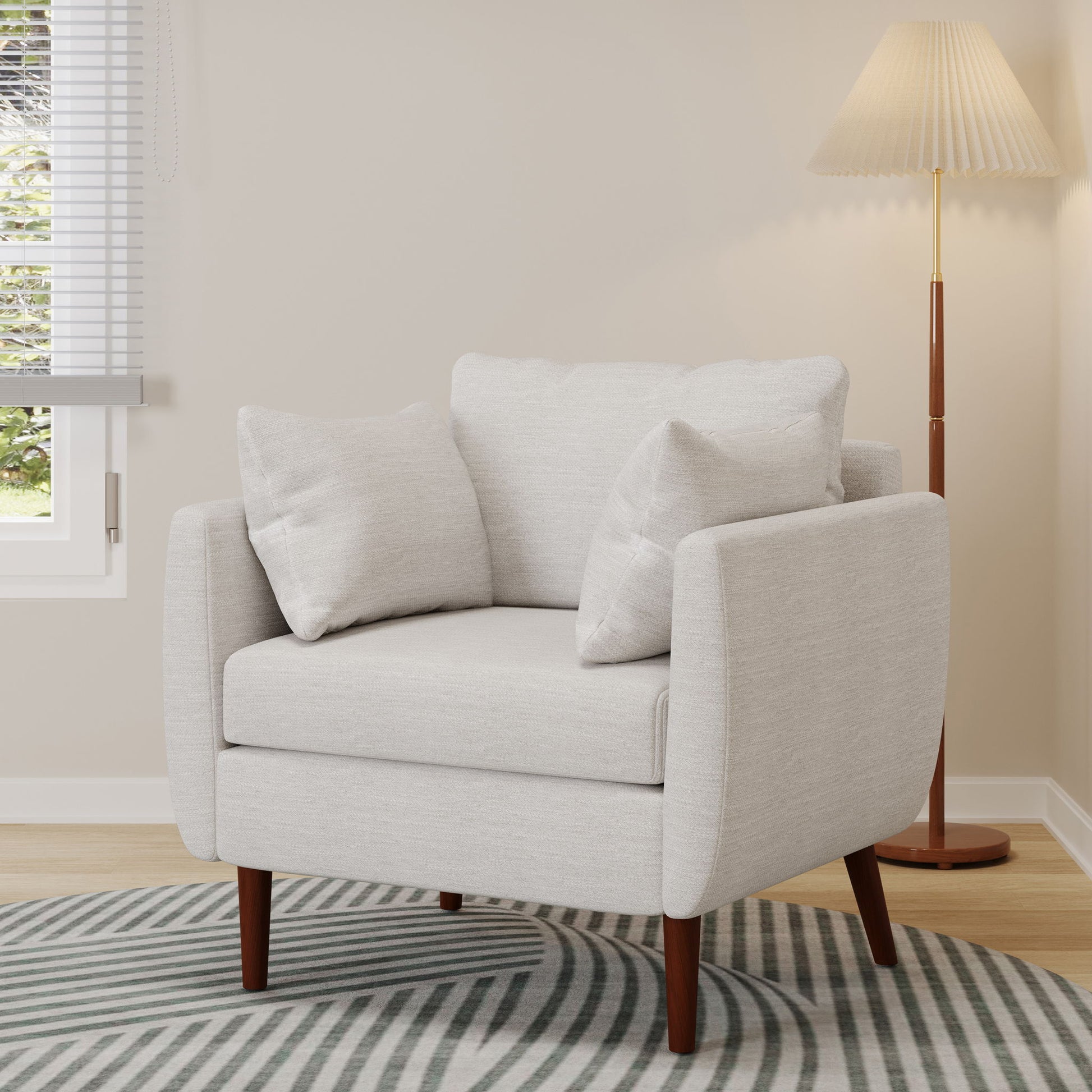 The Luminate Upholstered Fabric Club Chair with tapered legs and 2 accent pillows sits on a striped rug near a window, alongside a wooden floor lamp with a pleated shade in a softly lit room.