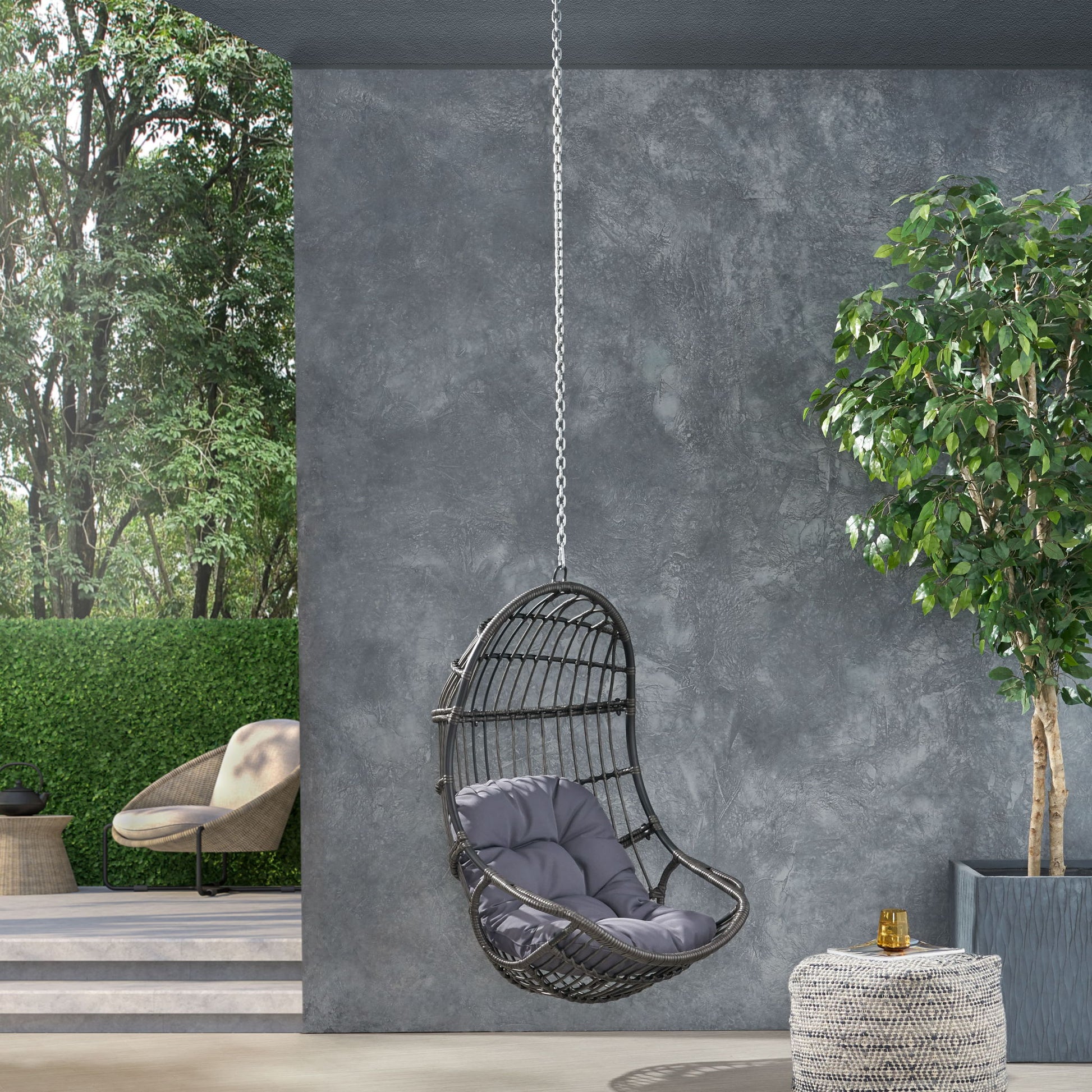 The Rustic Hanging Basket Chair With Cushion, in black with gray cushions, hangs by a chain on a modern patio. A potted tree, small table, and another chair sit nearby, surrounded by greenery and trees.