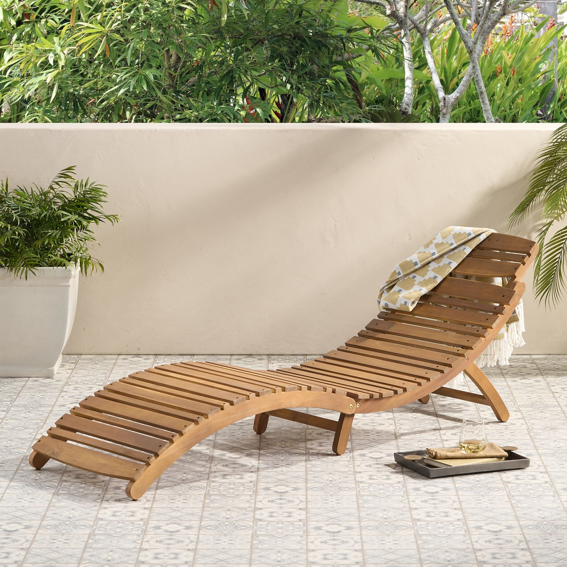 The Lahaina - Wood Foldable Chaise Lounge with a yellow-patterned towel is on a tiled patio beside a potted plant and tray with wine glass and book, set against lush green foliage.