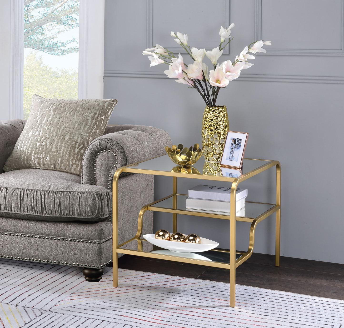 A modern living room corner with a gray tufted sofa and the Astrid Mirrored Table, featuring a gold vase of white flowers, a photo frame, and decorative objects on its lower shelves, set against a gray wall.