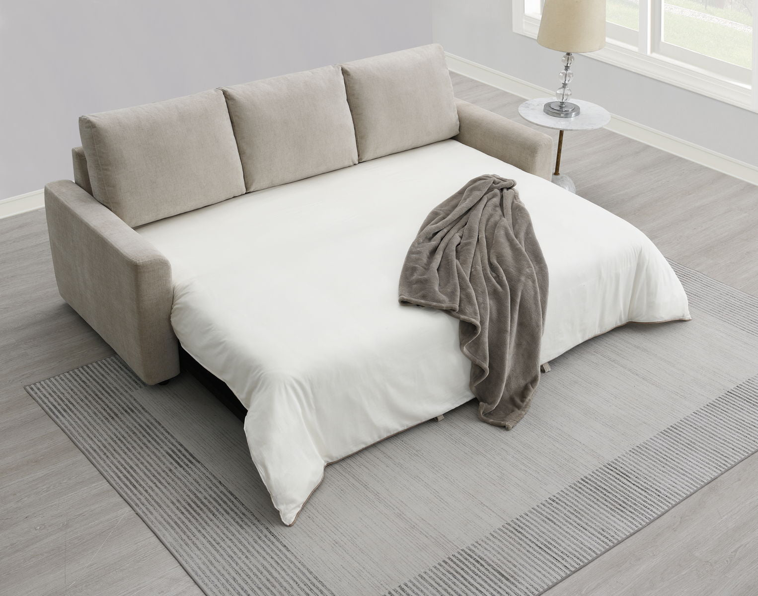 The Leira Linen Pull Out Sleeper Sofa in beige, with a white mattress and gray throw, is unfolded on a light gray rug in a bright room. A round side table with a lamp sits by the window.