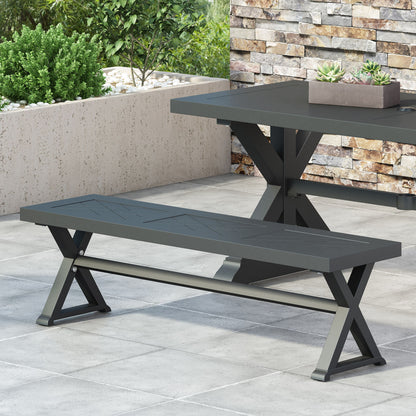 A sleek patio features the Modern Aluminum Outdoor Dining Bench With X Base and a matching table, both with X-shaped legs, set on gray tiles against a stone wall with green plants in the background.