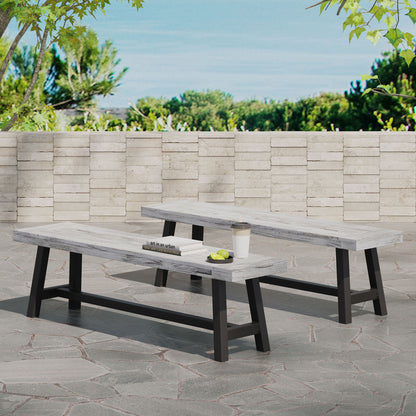 Two Tranquilizer outdoor acacia wood benches with slat panel design feature light wooden tops and black legs on a stone patio. A book, cup, and plate with fruit rest on one bench, set against a light stone wall and green trees.