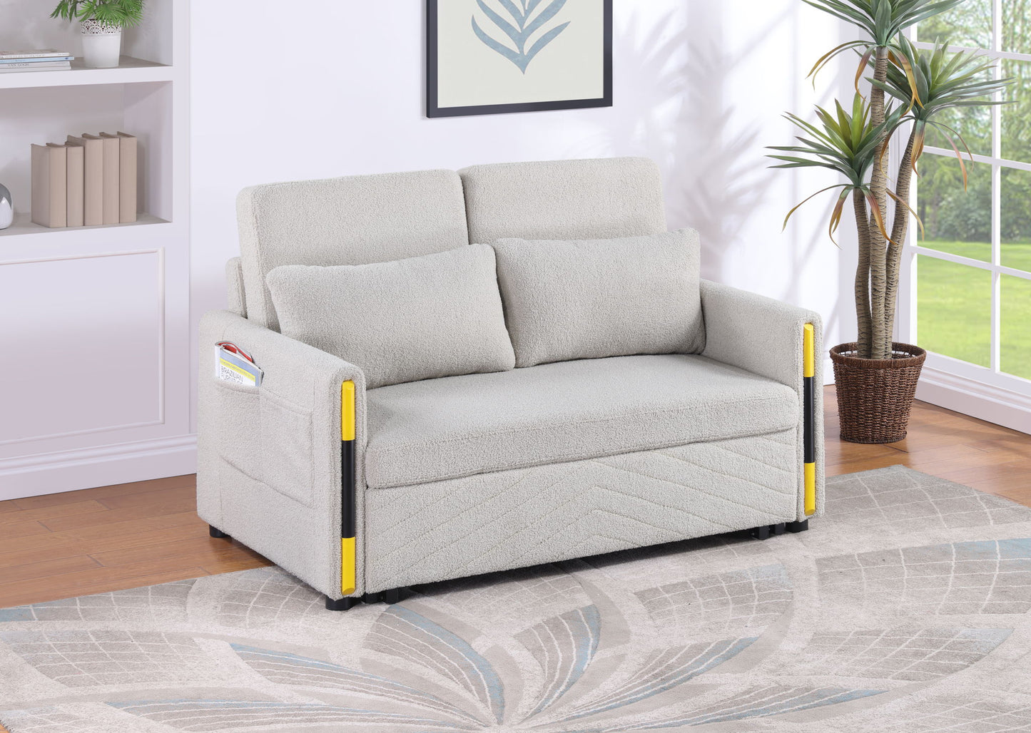 The Percy - Teddy Velvet Sleeper Loveseat features light gray upholstery, two cushions, yellow plastic armrest guards, a side magazine pocket, and sits on a patterned rug in a bright, modern living room.