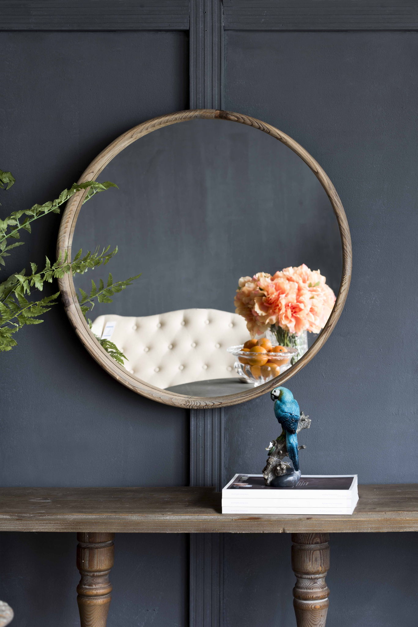 A Round Wood Mirror, Wall Mounted Mirror Home Decor For Bathroom Living Room, reflects a tufted cream chair and peach flowers. In front, a wooden table holds stacked books and a blue bird figurine. Green leaves frame the mirror’s left side.