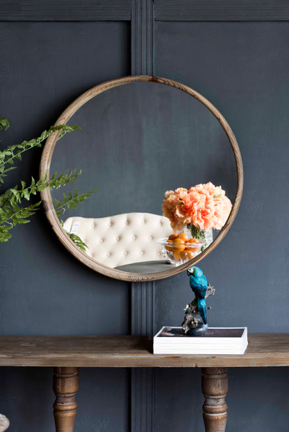 A Round Wood Mirror, Wall Mounted Mirror Home Decor For Bathroom Living Room, reflects a tufted cream chair and peach flowers. In front, a wooden table holds stacked books and a blue bird figurine. Green leaves frame the mirror’s left side.