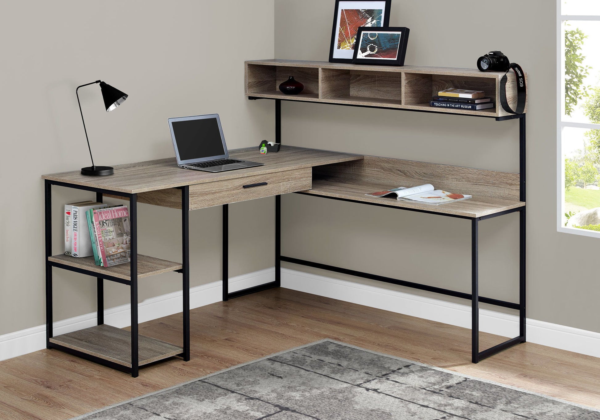 The Computer Desk, Home Office, Corner L Shape features storage drawers and a modern design, shown with a laptop, desk lamp, magazines, camera, and decor on shelves in a bright room with a window and gray rug.