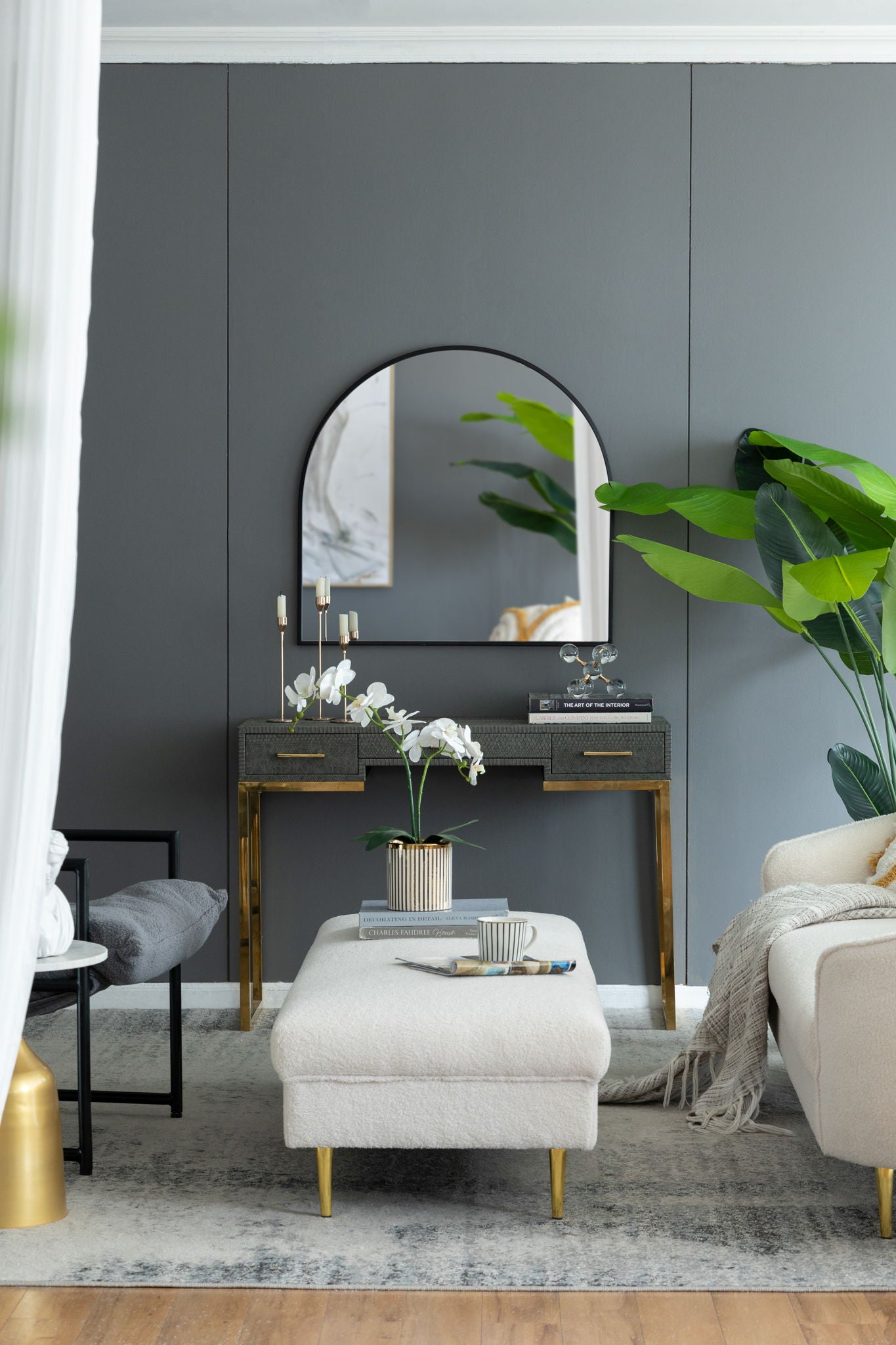 A modern living room with gray walls, a console table with gold accents, an Arched Mirror With Metal Frame for the wall, potted plants, candles, a light ottoman topped with a striped mug and saucer, a cream sofa, and stylish decor.