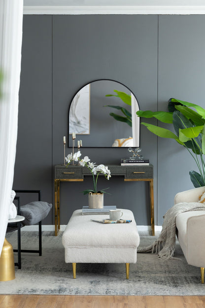 A modern living room with gray walls, a console table with gold accents, an Arched Mirror With Metal Frame for the wall, potted plants, candles, a light ottoman topped with a striped mug and saucer, a cream sofa, and stylish decor.