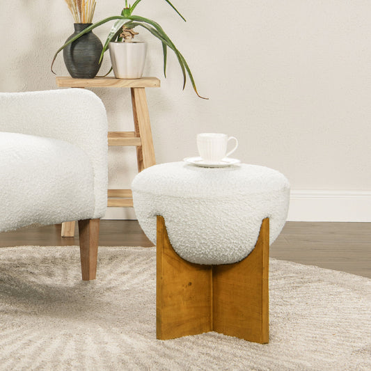 A modern living room showcases the Bali - Round Upholstered Ottoman with a wooden base, used as a side table for a white teacup and saucer, placed next to a white sofa and a wooden shelf with plants.