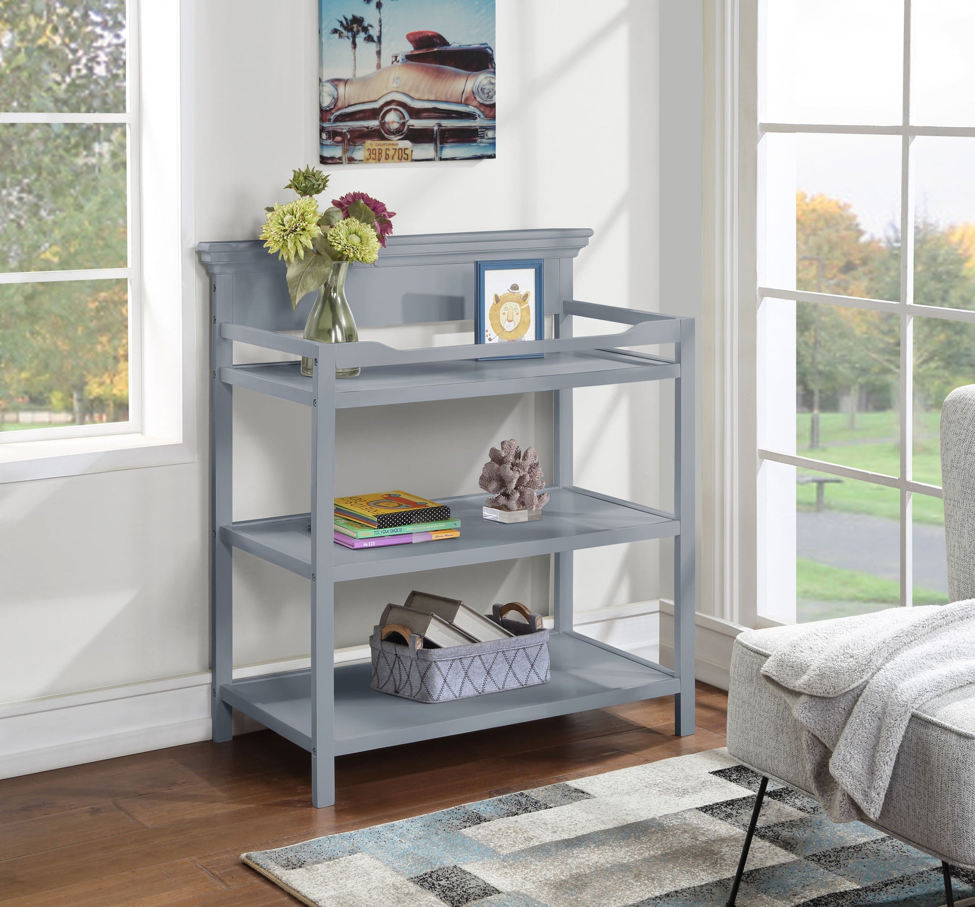 A gray Universal Changing Table stands by large windows, displaying vases with flowers, books, a framed picture, a decorative sculpture, and a storage basket. Above it hangs a car painting; part of a sofa is also visible nearby.