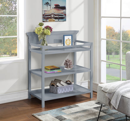 A gray Universal Changing Table stands by large windows, displaying vases with flowers, books, a framed picture, a decorative sculpture, and a storage basket. Above it hangs a car painting; part of a sofa is also visible nearby.