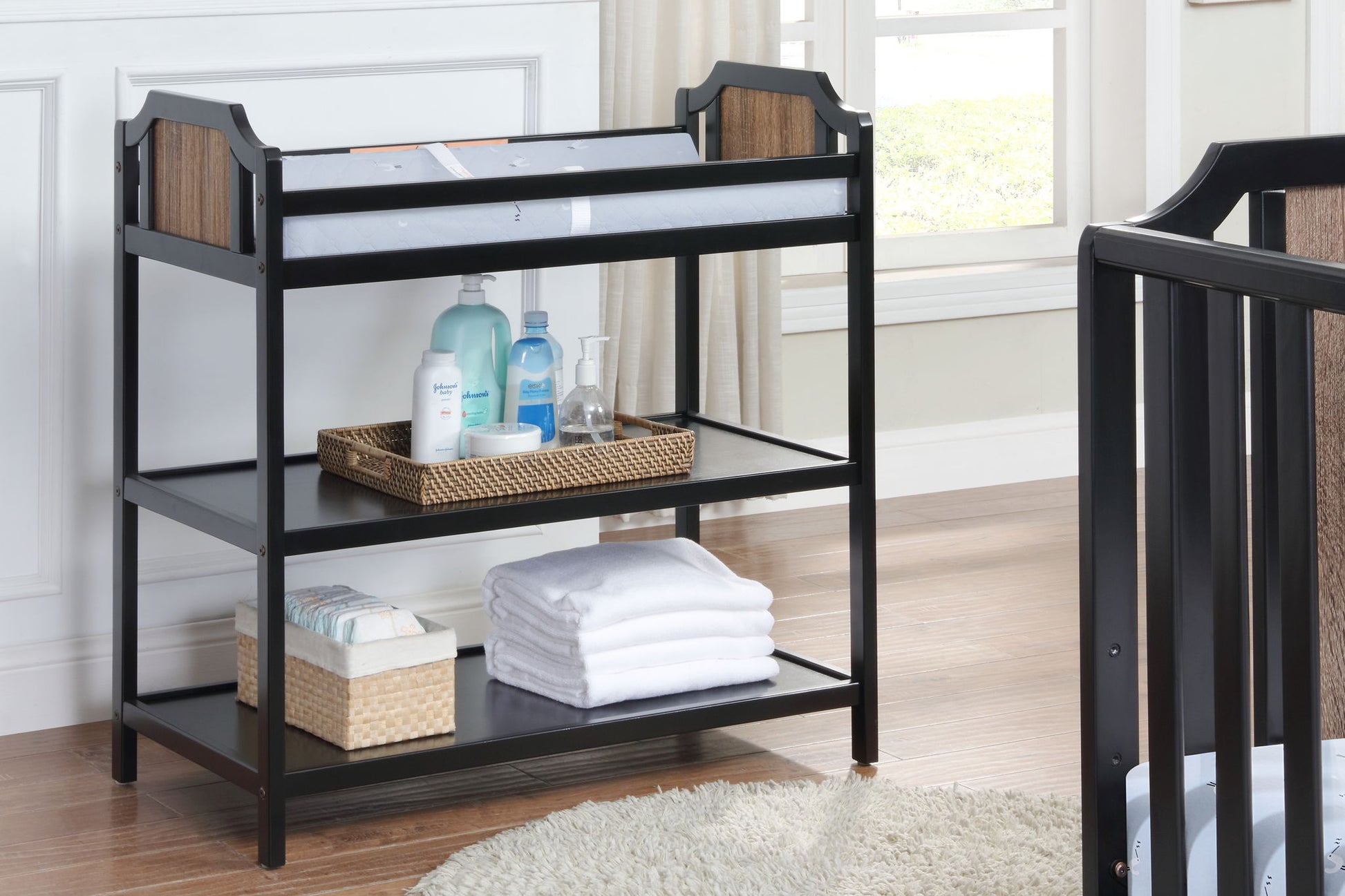 The Brees - Changing Table in black and wood features two shelves for items like folded white towels, baby wipes, and lotions, perfect for organizing baby care essentials in a bright nursery with light wood flooring.