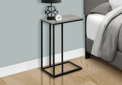 The Accent Table, C-Shaped, Space-Saving Functionality features a gray wood top and black metal frame beside a bed, holding a geometric black lamp and small white clock. Light wood floor and light blue wall complete the setting.